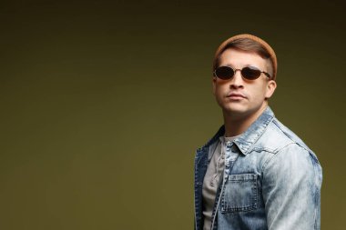 Moda modeli. Şapkalı, güneş gözlüklü ve ceketli, koyu yeşil arkaplanda yakışıklı bir adam.