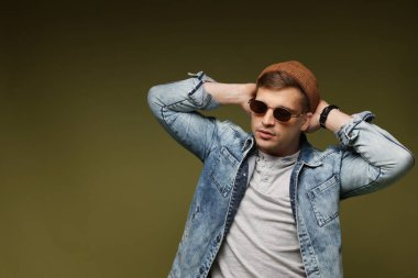 Moda modeli. Şapkalı, güneş gözlüklü, ceketli, koyu yeşil arkaplanda poz veren yakışıklı bir adam.