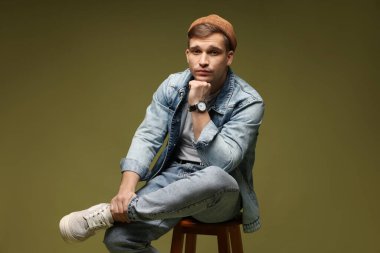 Moda modeli. Şapkalı ve ceketli yakışıklı adam, koyu yeşil arka planda dışkı üzerinde poz veriyor.