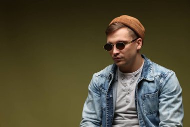 Moda modeli. Şapkalı, güneş gözlüklü ve ceketli, koyu yeşil arkaplanda yakışıklı bir adam.