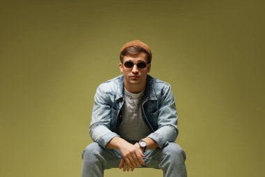 Moda modeli. Şapkalı, güneş gözlüklü ve ceketli yakışıklı adam koyu yeşil eğimli arka plana karşı sandalyede poz veriyor.