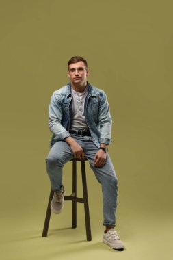 Moda modeli. Yeşil arka planda taburede poz veren gözlüklü ve ceketli yakışıklı adam.