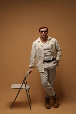 Moda modeli. Şapkalı, güneş gözlüklü, bej arka planda sandalyesi olan yakışıklı adam.