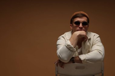 Moda modeli. Şapkalı ve güneş gözlüklü yakışıklı, kahverengi arka planda sandalyede poz veriyor, mesaj için yer var.