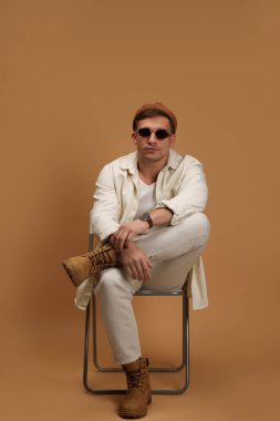 Moda modeli. Şapkalı ve güneş gözlüklü yakışıklı bej arka planda sandalyede poz veriyor.