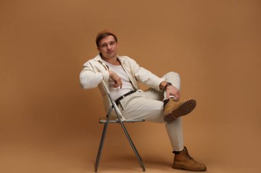 Moda modeli. Sandalyede bej arka planda poz veren şapkalı yakışıklı bir adam.