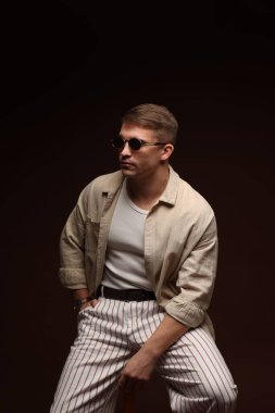 Moda modeli. Siyah arka planda taburede poz veren güneş gözlüklü yakışıklı bir adam.