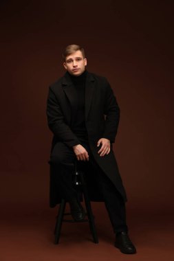 Moda modeli. Koyu kahverengi arka planda taburede poz veren yakışıklı bir adam.