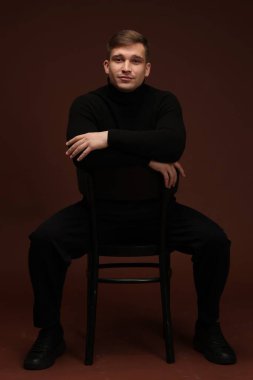 Moda modeli. Koyu kahverengi arka planda sandalyede poz veren yakışıklı bir adam.