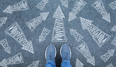 Spor ayakkabılı bir adam yolda duruyor, oklar farklı yönlere bakıyor, üst manzara. Yolculuğuna başla.