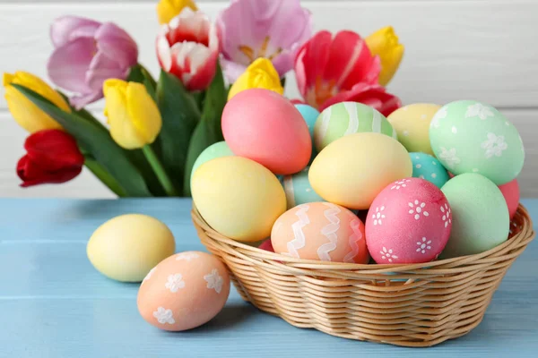 Hasır sepetin içine boyanmış Paskalya yumurtaları açık mavi ahşap masa ve kapalı alanda güzel laleler. Yakın plan.