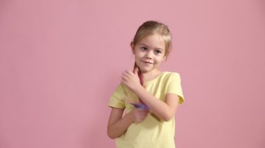 Renkli saç eklentileri olan küçük bir kız pembe arka planda baş parmaklarını gösteriyor.