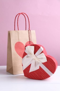 Kağıt torba ve kalp şeklinde hediye paketi beyaz masada, pembe arka planda, yakın plan. Sevgililer Günü