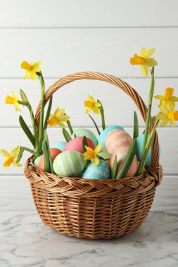 Paskalya yumurtaları ve hasır sepetin içindeki güzel nergisler beyaz mermer masanın üzerinde, yakın plan.