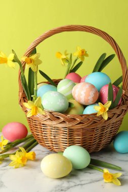 Boyanmış Paskalya yumurtaları ve hasır sepetin içindeki güzel nergisler açık yeşil arka planda beyaz mermer masa üzerinde, yakın plan