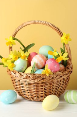 Boyanmış Paskalya yumurtaları ve hasır sepetin içindeki güzel nergisler bej arka planda beyaz ahşap masa üzerinde, yakın plan