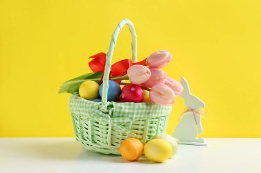 Hasır sepette güzel lalelerle boyanmış Paskalya yumurtaları ve sarı arka planda beyaz masa üzerinde dekoratif tavşancık.