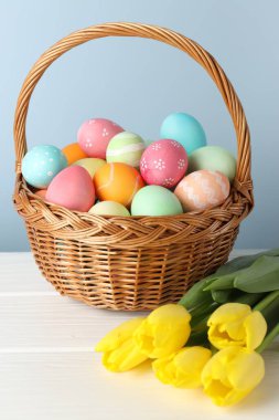 Hasır sepete boyanmış Paskalya yumurtaları ve beyaz ahşap masada açık mavi arka planda güzel laleler.