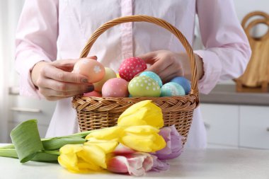 Paskalya sepetinde yumurta boyalı bir kadın ve beyaz masa içinde laleler, yakın plan.