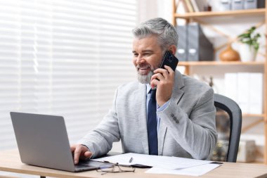 Takım elbiseli gülümseyen işadamı akıllı telefonla konuşuyor ve ofiste dizüstü bilgisayarla çalışıyor.