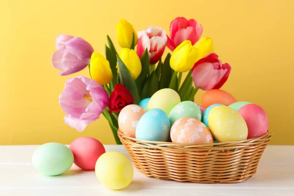 Hasır sepetin içine boyanmış Paskalya yumurtaları beyaz ahşap masa üzerinde ve sarı arka planda güzel laleler, yakın plan.