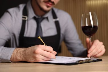 Profesyonel şarap garsonu, kırmızı şarap kadehiyle ahşap masada tadımlık kartı dolduruyor.