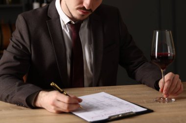 Profesyonel şarap garsonu, kırmızı şarap kadehiyle ahşap masada tadımlık kartı dolduruyor.