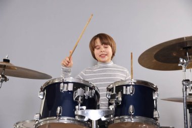 Açık gri arka planda bagetleri ve davulları olan küçük bir çocuk.