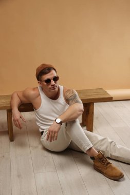 Moda modeli. Şapkalı ve güneş gözlüklü yakışıklı adam. Yerde poz veriyor. Bankta bej arka plana yaslanıyor.