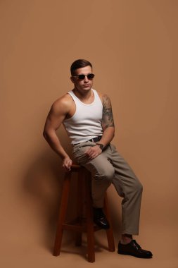 Moda modeli. Güneş gözlüklü yakışıklı adam bej arka planda taburede poz veriyor.