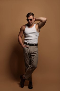 Moda modeli. Güneş gözlüklü yakışıklı adam bej arka planda poz veriyor.