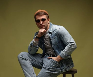 Moda modeli. Şapkalı, güneş gözlüklü ve ceketli yakışıklı adam koyu yeşil renkli arka plana karşı dışkı üzerinde poz veriyor.
