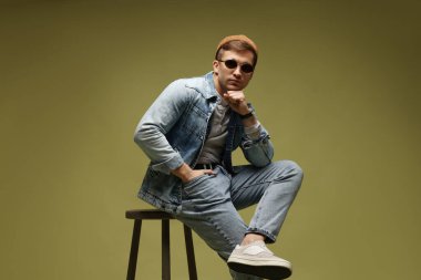 Moda modeli. Şapkalı, güneş gözlüklü ve ceketli yakışıklı adam koyu yeşil renkli arka plana karşı dışkı üzerinde poz veriyor.