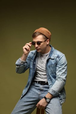 Moda modeli. Şapkalı, güneş gözlüklü ve ceketli yakışıklı adam koyu yeşil renkli arka plana karşı dışkı üzerinde poz veriyor.