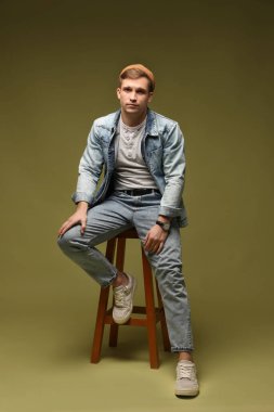 Moda modeli. Şapkalı ve ceketli yakışıklı adam, koyu yeşil arka planda dışkı üzerinde poz veriyor.