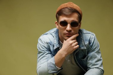 Moda modeli. Şapkalı, güneş gözlüklü ve ceketli yakışıklı adam yeşil arka planda, mesaj için yer var.
