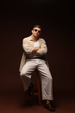 Moda modeli. Güneş gözlüklü yakışıklı, siyah arka planda kolları çapraz duran bir adam.