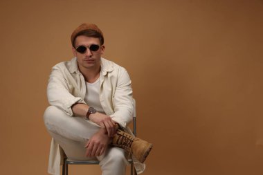 Moda modeli. Şapkalı ve güneş gözlüklü yakışıklı adam bej arka plana karşı sandalyede poz veriyor, mesaj için yer var.