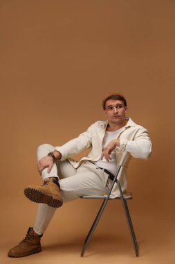 Moda modeli. Sandalyede bej arka planda poz veren şapkalı yakışıklı bir adam.