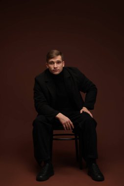 Moda modeli. Koyu kahverengi arka planda sandalyede poz veren yakışıklı bir adam.