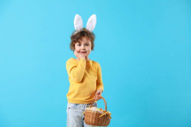 Paskalya kutlaması. Saç bantlı, tavşan kulaklı ve açık mavi arka planda boyalı yumurtalarla dolu sepeti olan şirin bir çocuk. Metin için boşluk