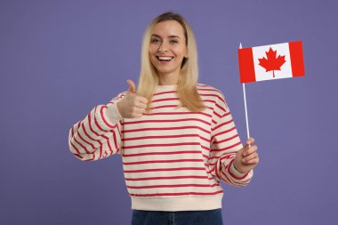 Kanada bayrağı taşıyan mutlu kadın pastel mor arka planda baş parmak gösteriyor.