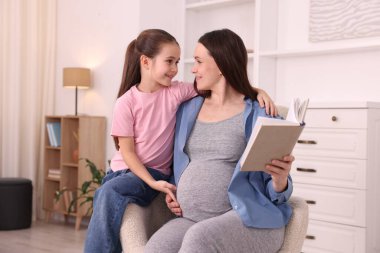 Hamile kadın ve kızı evdeki koltukta kitap okuyorlar.