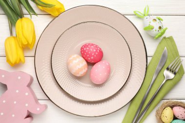 Paskalya kutlaması. Çatal bıçak, çiçek ve beyaz ahşap masaya boyanmış yumurtalarla şenlikli bir ortam.