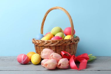 Hasır sepete boyanmış Paskalya yumurtaları ve açık mavi arka plana karşı gri ahşap masada güzel laleler.