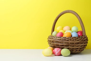 Hasır sepetin içine boyanmış Paskalya yumurtaları beyaz masada, sarı arka planda. Metin için boşluk