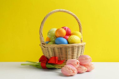Hasır sepette boyanmış Paskalya yumurtaları ve sarı arka planda beyaz masada güzel laleler.