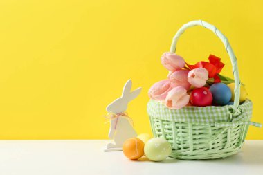Hasır sepette güzel lalelerle boyanmış Paskalya yumurtaları ve sarı arka planda beyaz masa üzerine dekoratif tavşancık. Metin için boşluk