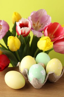 Ahşap masada karton içine boyanmış Paskalya yumurtaları ve sarı arka planda güzel laleler. Yakın plan.