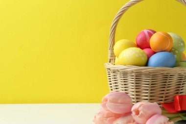 Hasır sepete boyanmış Paskalya yumurtaları ve sarı arka planda beyaz masada güzel laleler. Yakın plan. Metin için boşluk